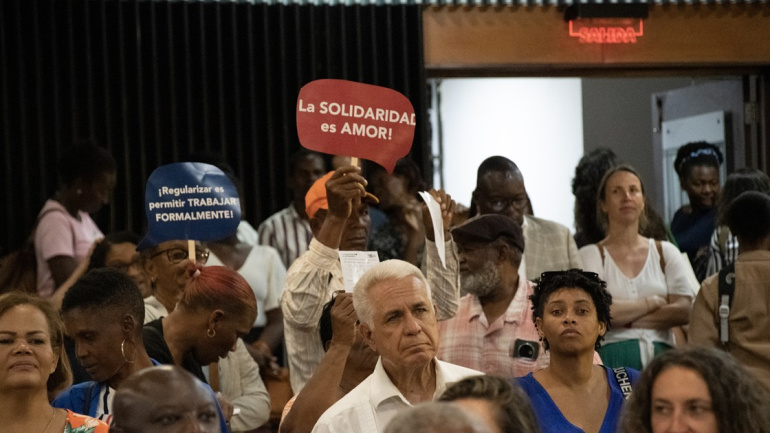 Personas asistentes a un evento del Colectivo Migración y Derechos Humanos sostienen carteles con los mensajes 'Regularizar es permitir trabajar formalmente' y 'La solidaridad es amor'.